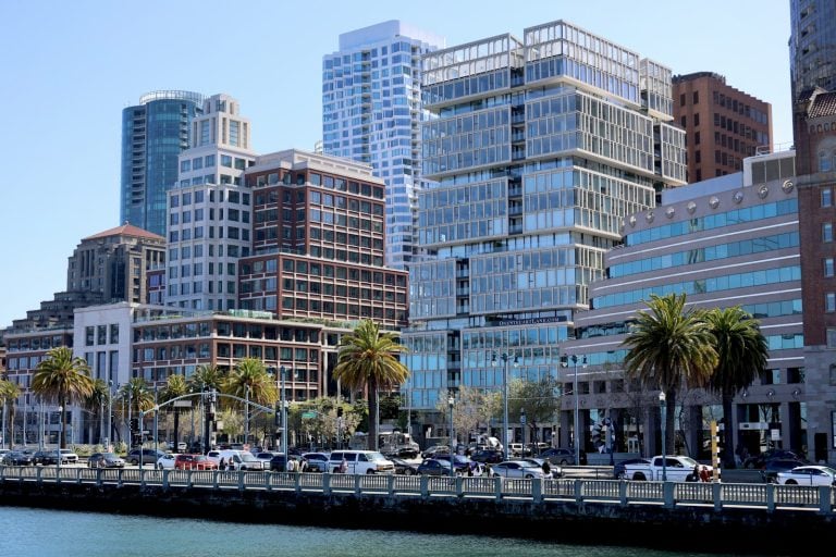 A cityscape features modern buildings, vehicles, and palm trees.